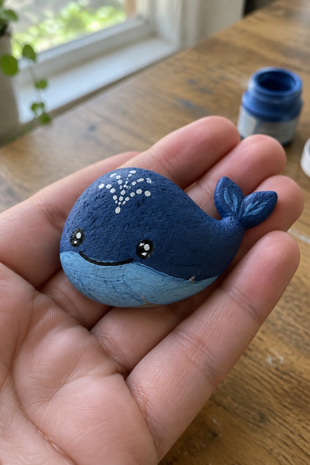 Hand holding small blue painted whale rock with smiling face on wooden table