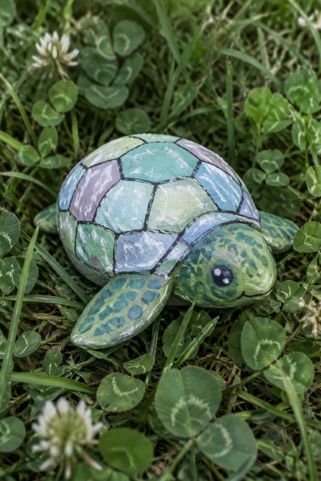 Painted rock sea turtle in green, blue, and purple resting among clover and grass