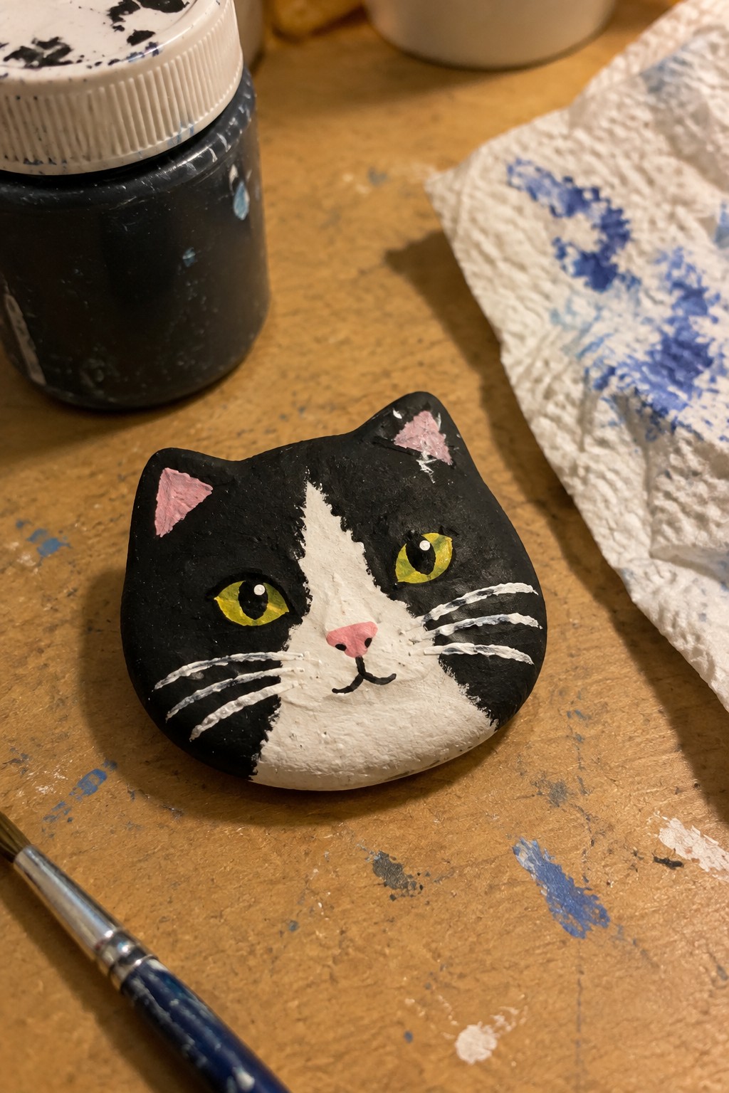 Black and white painted rock cat with yellow eyes beside paint jar and brush