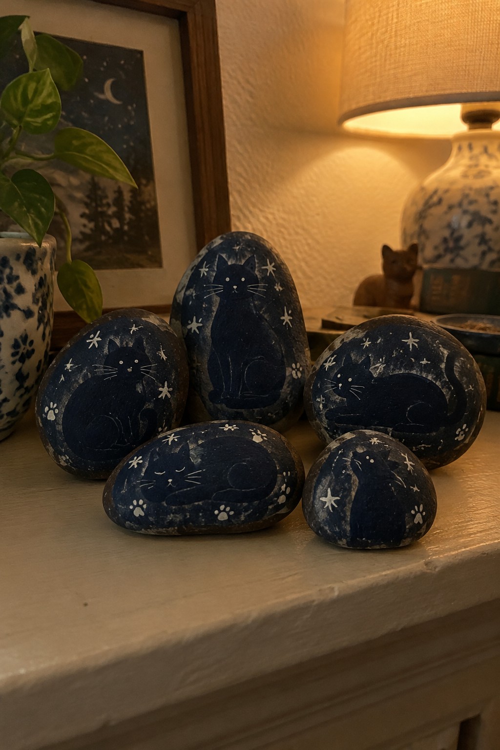 Five painted rocks with black cats, stars and paw prints on a cozy shelf.