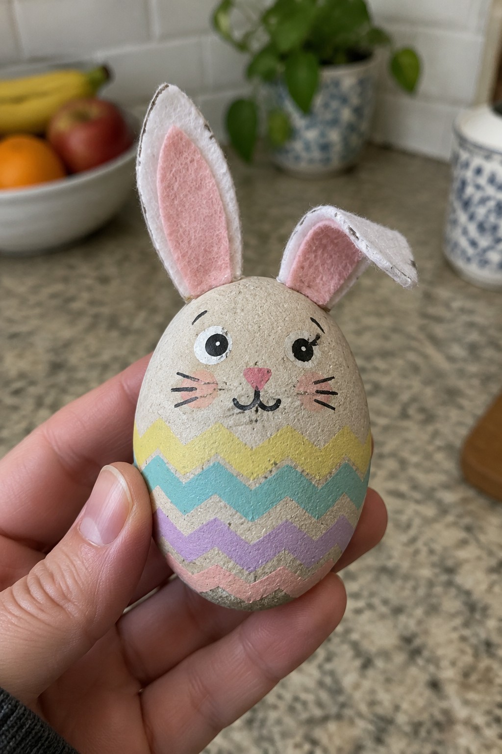 Hand holding a painted bunny Easter egg with pink felt ears and pastel chevrons.