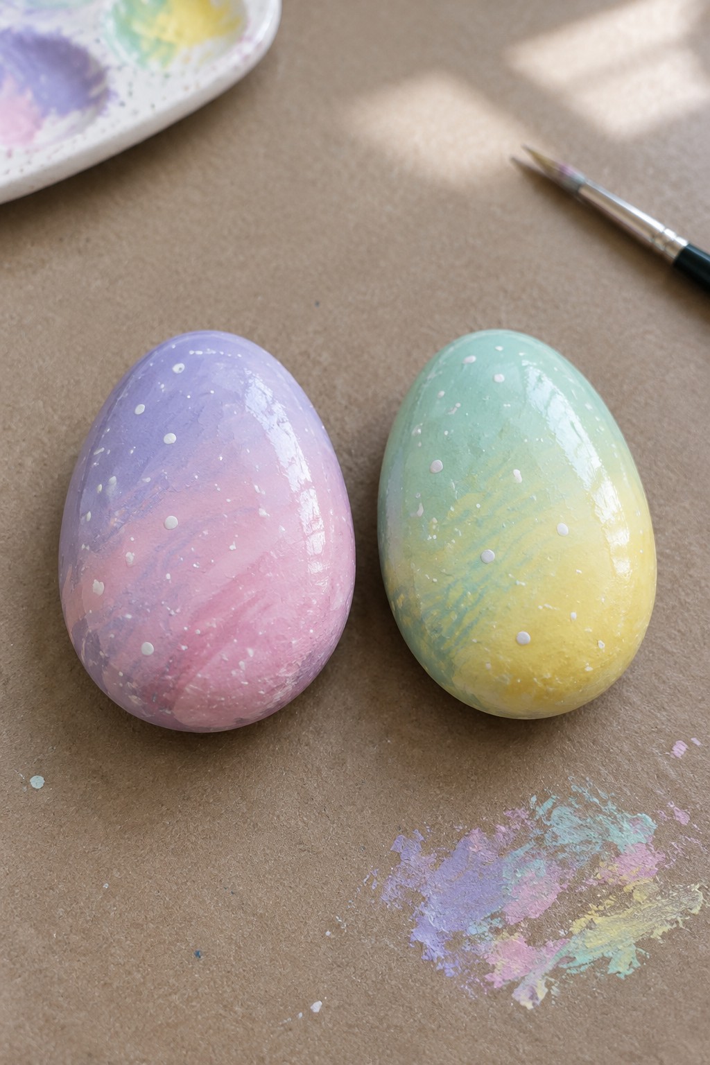Two glossy marbled pastel eggs beside a paint palette and brush on brown paper.
