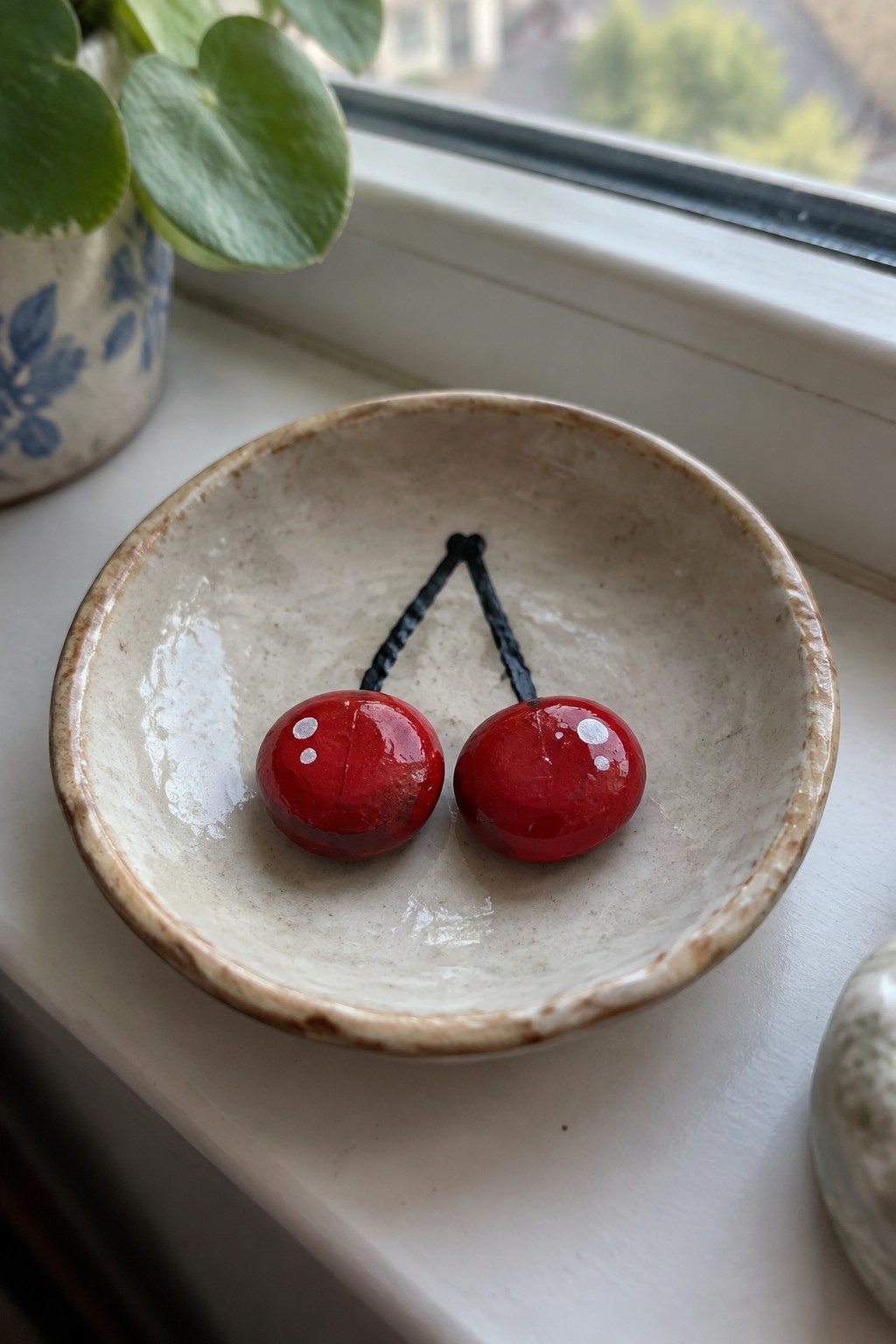 Two glossy red cherries with black stems in a speckled ceramic bowl on a windowsill