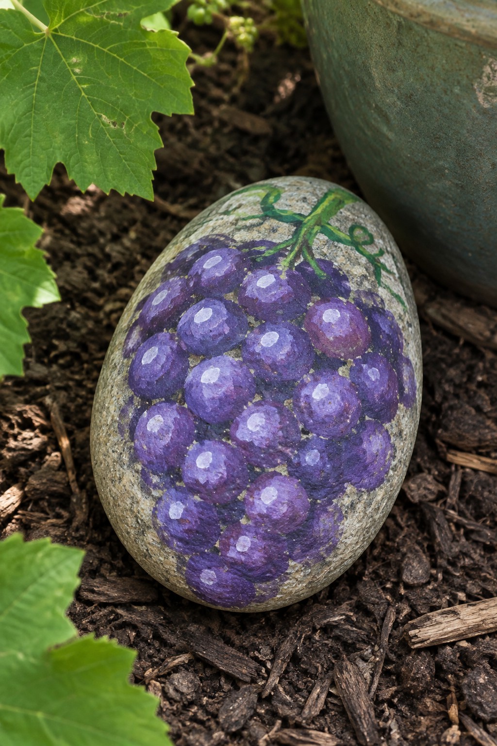 Stone painted with realistic purple grapes and green vine on garden soil.