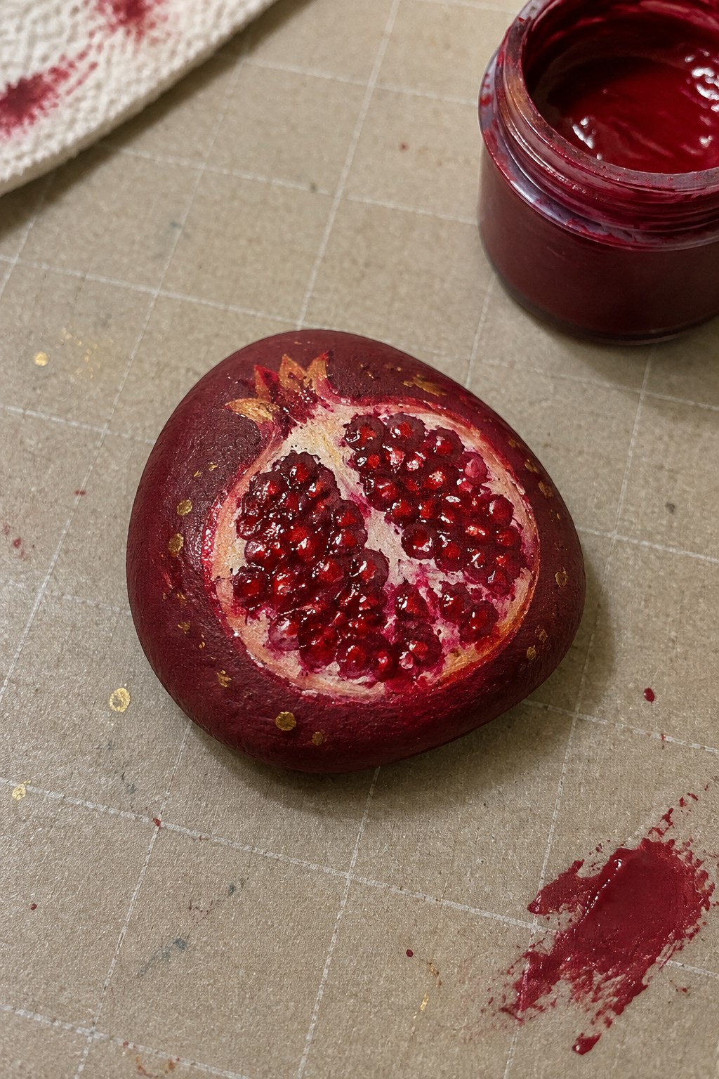 Painted pomegranate rock beside open red paint jar on grid mat with gold accents.