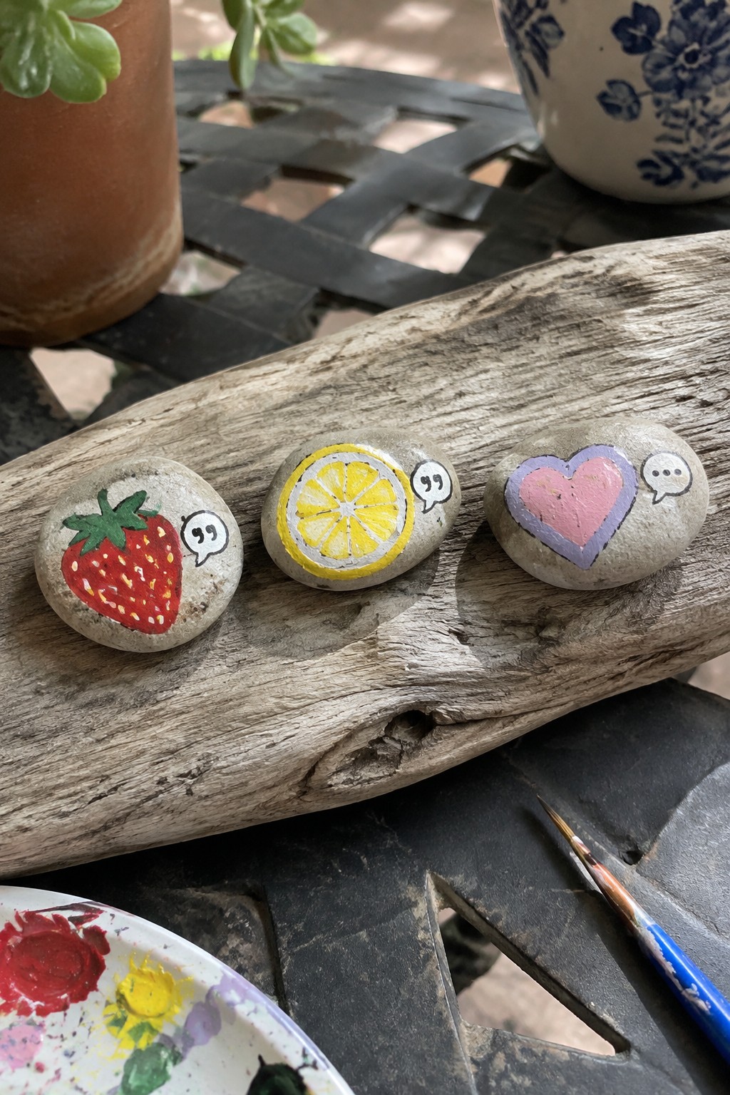 Three painted rocks with strawberry, lemon slice, and heart on weathered wood beside palette.