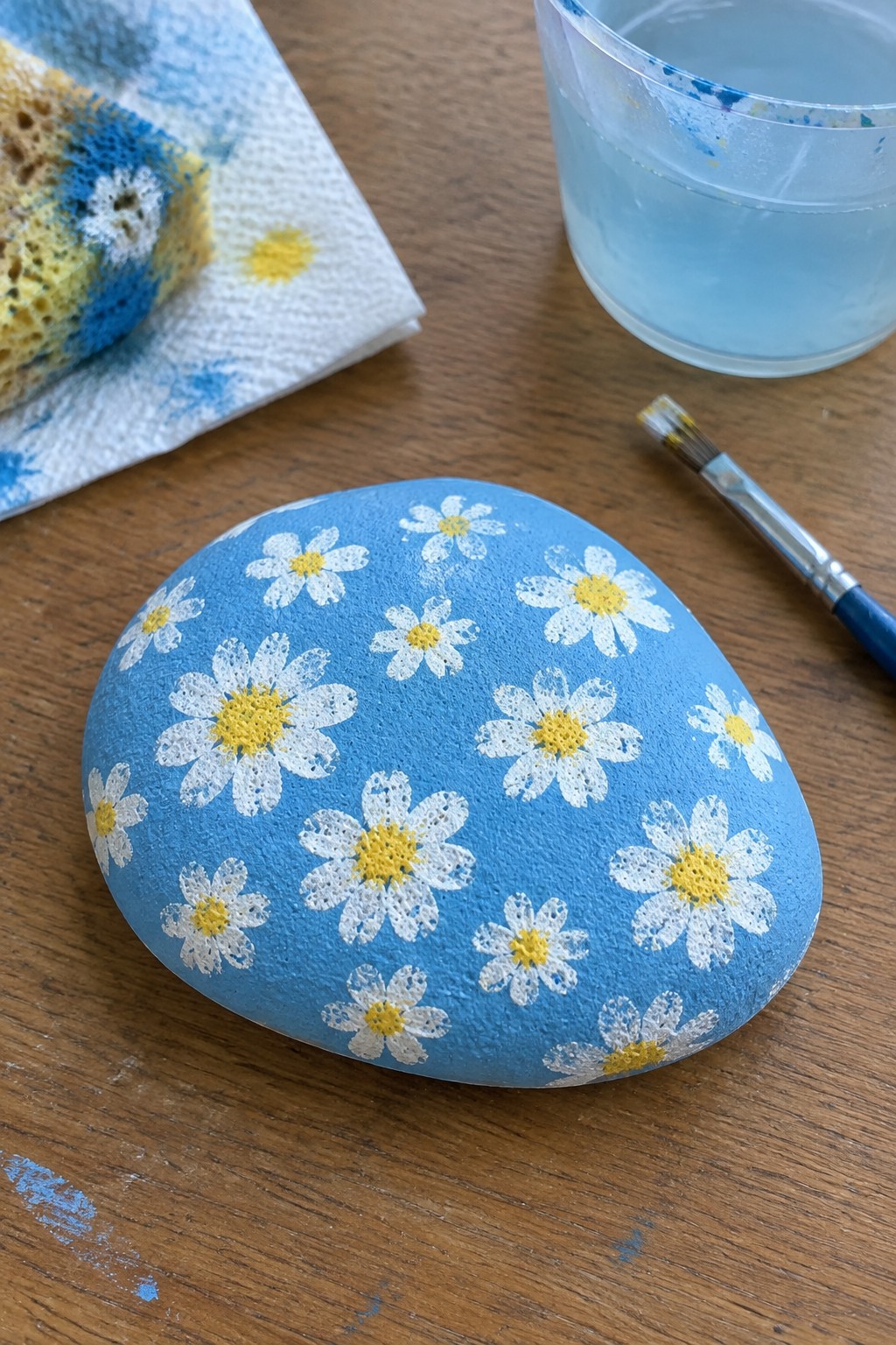 Blue rock with white daisies and yellow centers beside paintbrush on wooden table