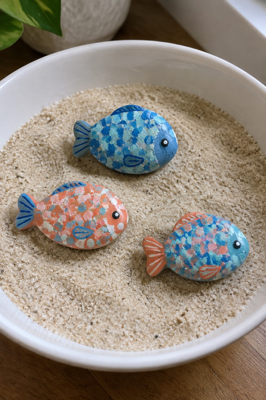 Three colorful painted fish stones nestled in sand inside white bowl