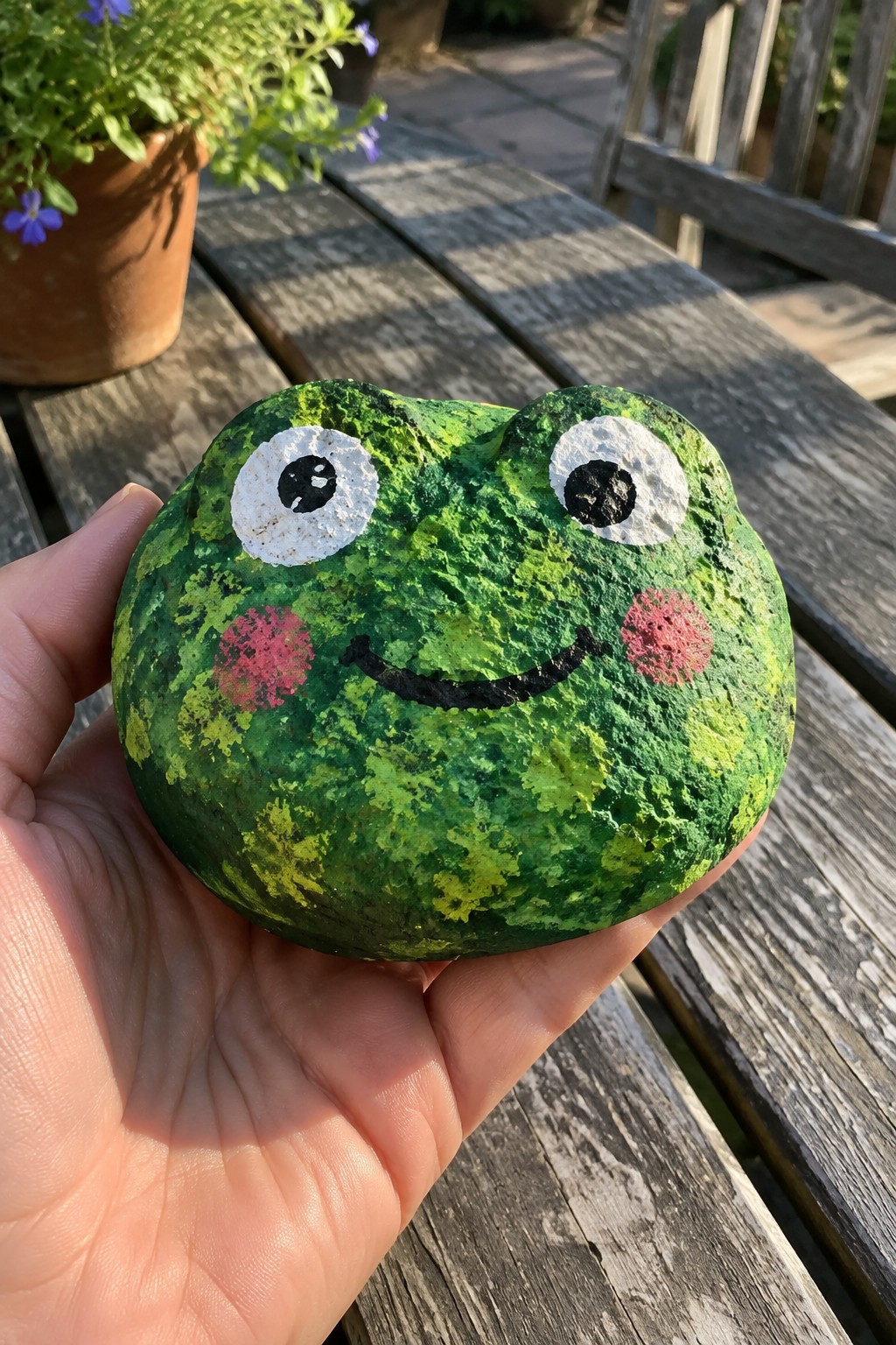 Hand holding green painted frog rock with white eyes and pink cheeks outdoors