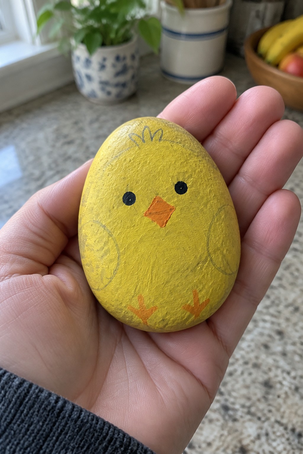 Hand holding yellow painted rock chick with black eyes, orange beak and feet.