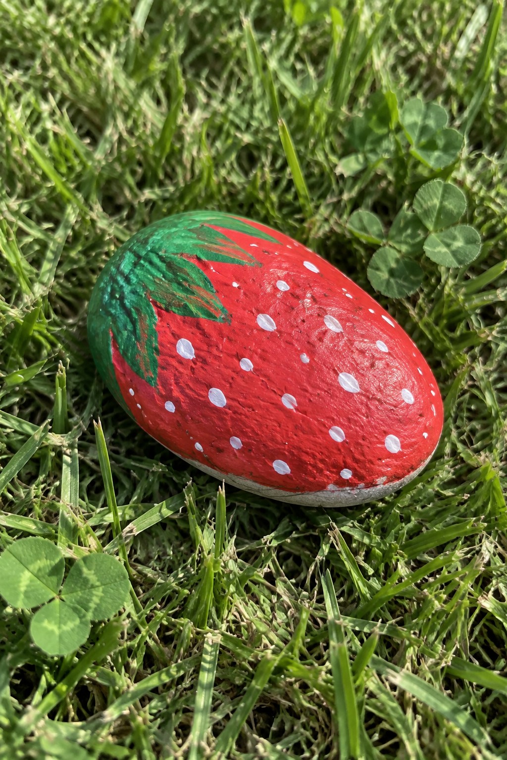 Red strawberry-painted rock with white dots and green leaves on grass