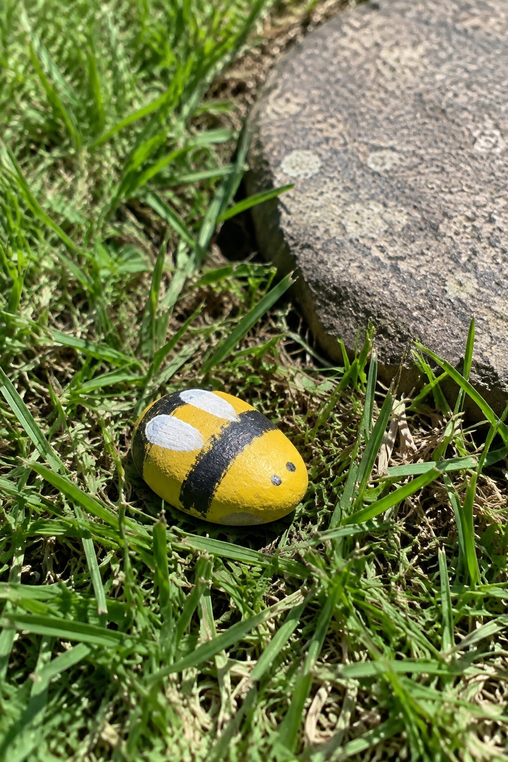 Yellow rock painted as bumblebee with black stripes, white wings, and eyes on grass