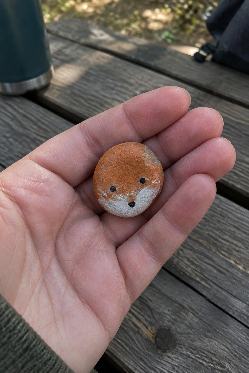 Hand holding small orange-white pebble painted as fox face on wood table