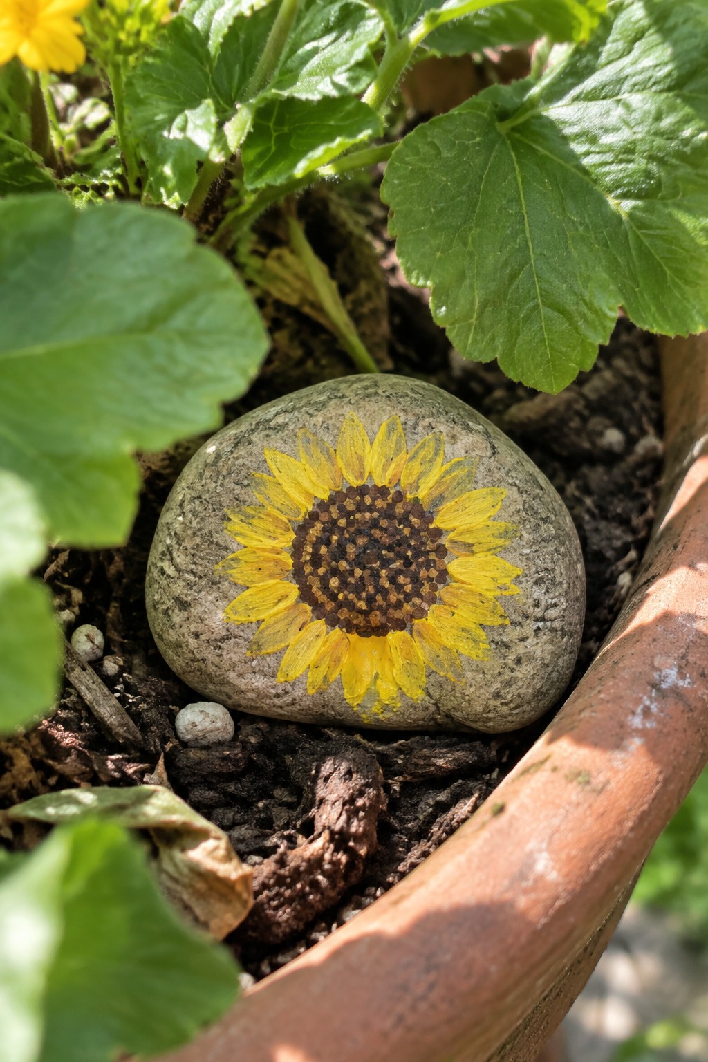 Sunflower painted on round stone in terracotta pot amid green leaves and soil