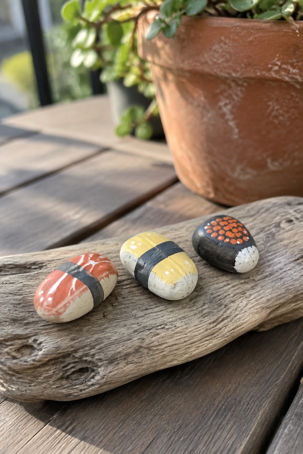 Three sushi-themed painted rocks on weathered driftwood near a terracotta plant pot.