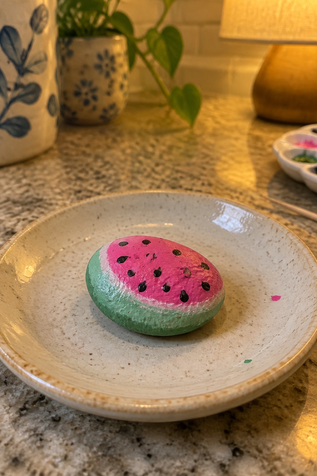Watermelon-painted rock with pink flesh and black seeds on speckled plate.