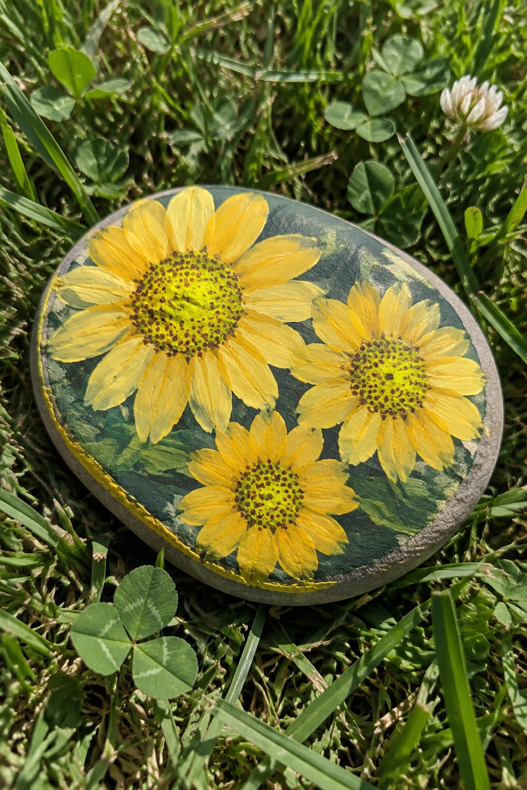 Oval painted rock with three yellow sunflowers on green leafy background in grass