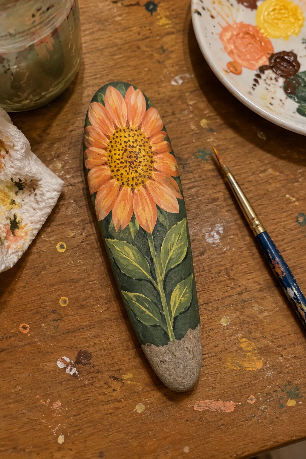 Painted sunflower on rock with orange petals, yellow center, green leaves on wooden table