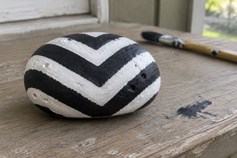 17 Cool Rock Painting Ideas Featuring Bold Geometric Designs A round stone painted with black and white chevron stripes sits on a wooden surface next to a paintbrush.
