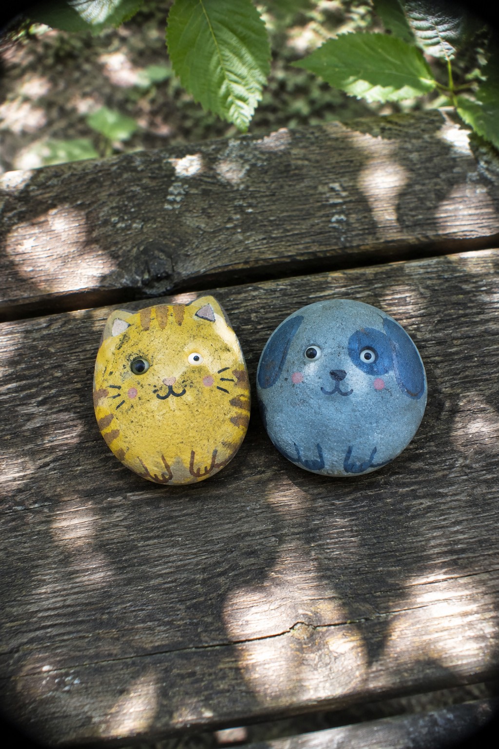 Two painted rocks rest on a wooden surface, one yellow with a cat face and one blue with a dog face, near green leaves.