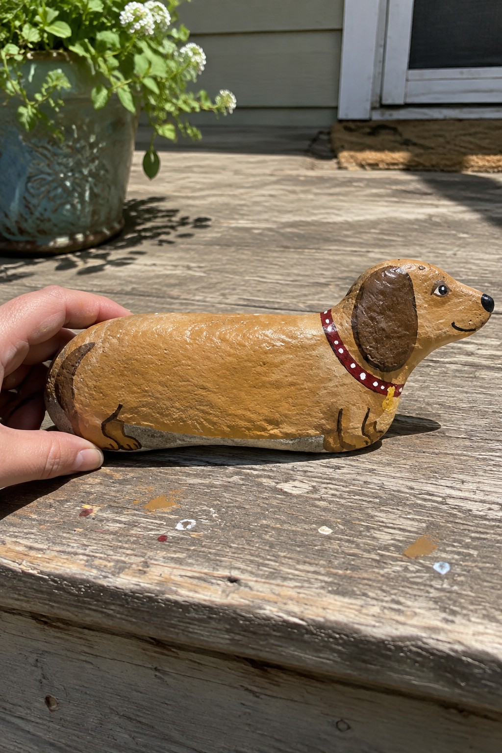 Hand holding tan painted dachshund rock with red dotted collar on wooden deck