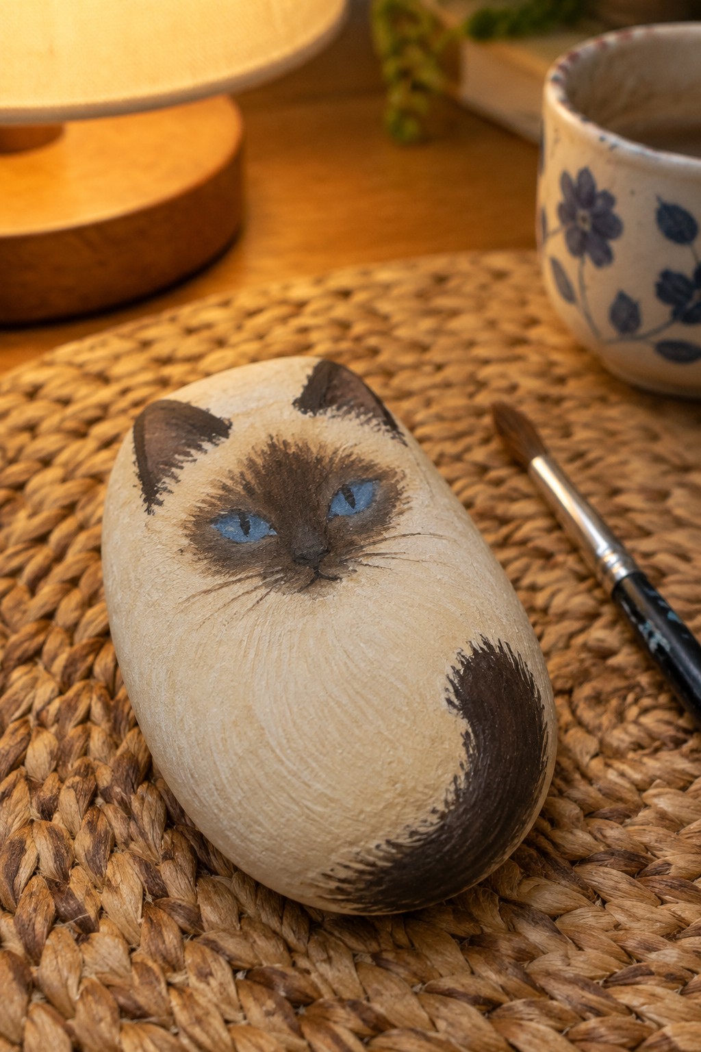 Rock painted as Siamese cat with blue eyes on woven mat beside paintbrush