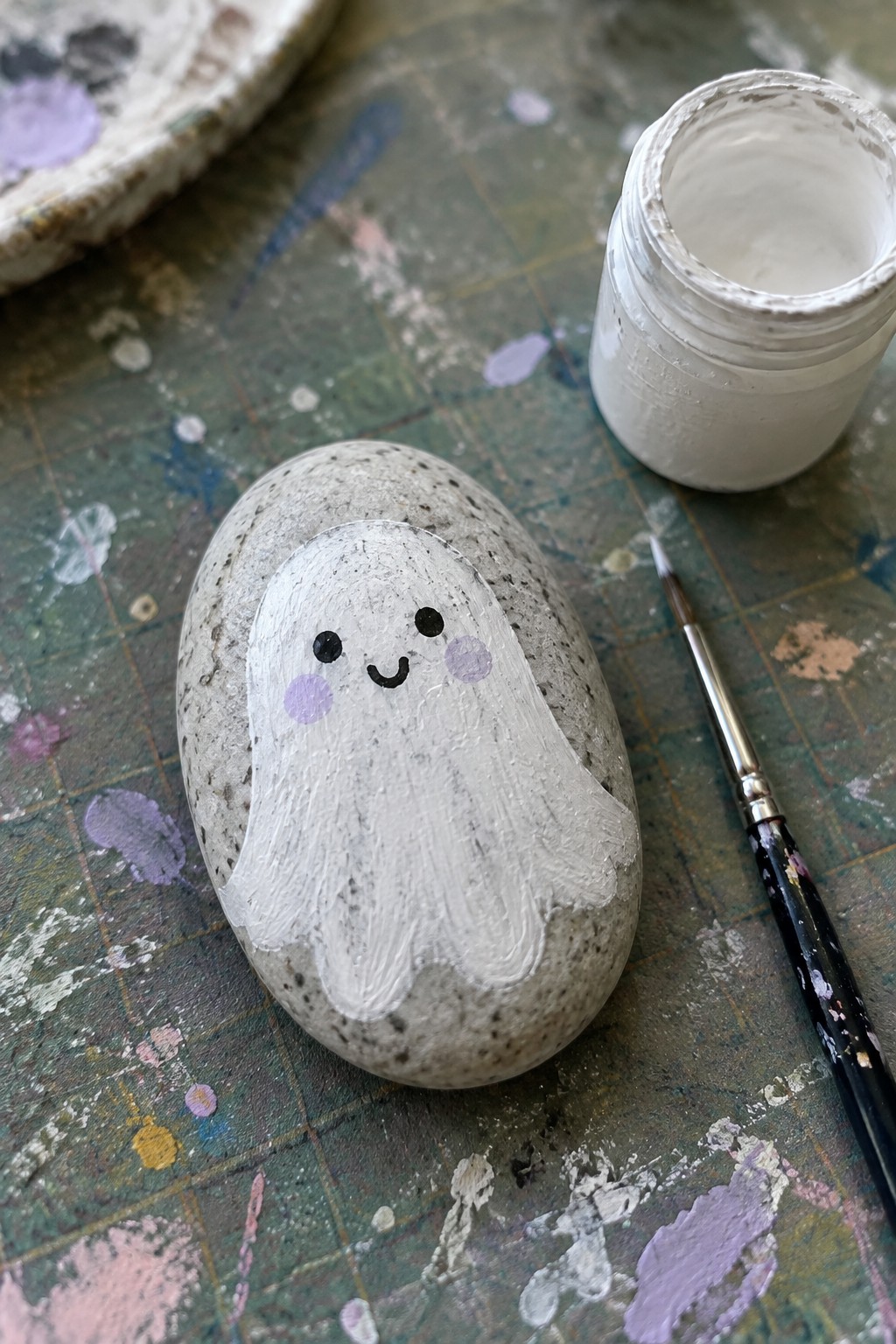Painted ghost rock with black eyes, purple cheeks beside paint jar and brush