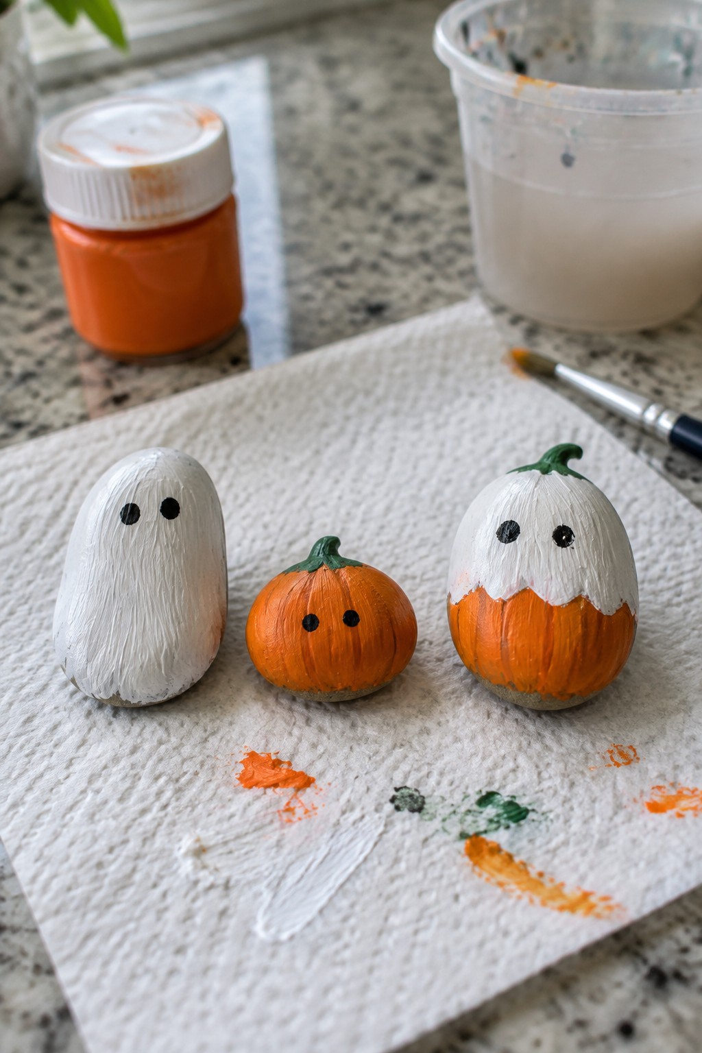 Three painted rocks as ghost and pumpkins on paper towel with orange paint jar and brush