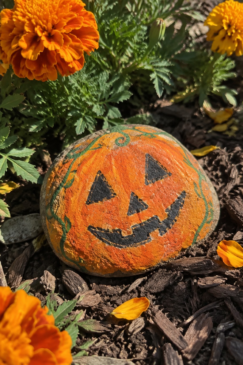 Jack-o'-lantern painted rock amid orange marigolds and garden mulch
