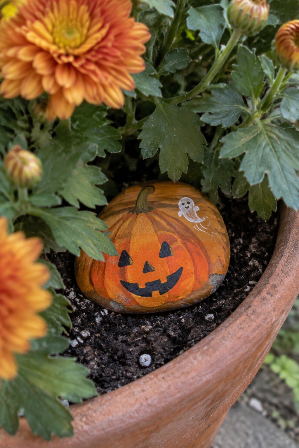 Orange jack-o'-lantern painted rock with ghost in potted soil among flowers