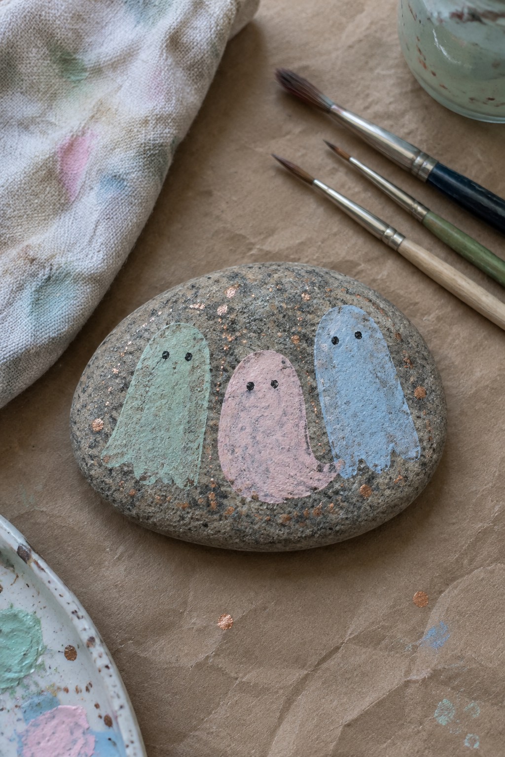 Painted stone with three pastel ghosts beside paintbrushes, palette, and cloth on paper