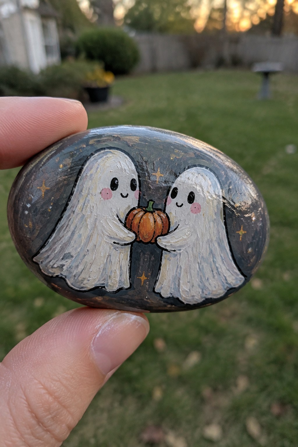 Hand holding painted rock of two smiling ghosts sharing orange pumpkin with stars