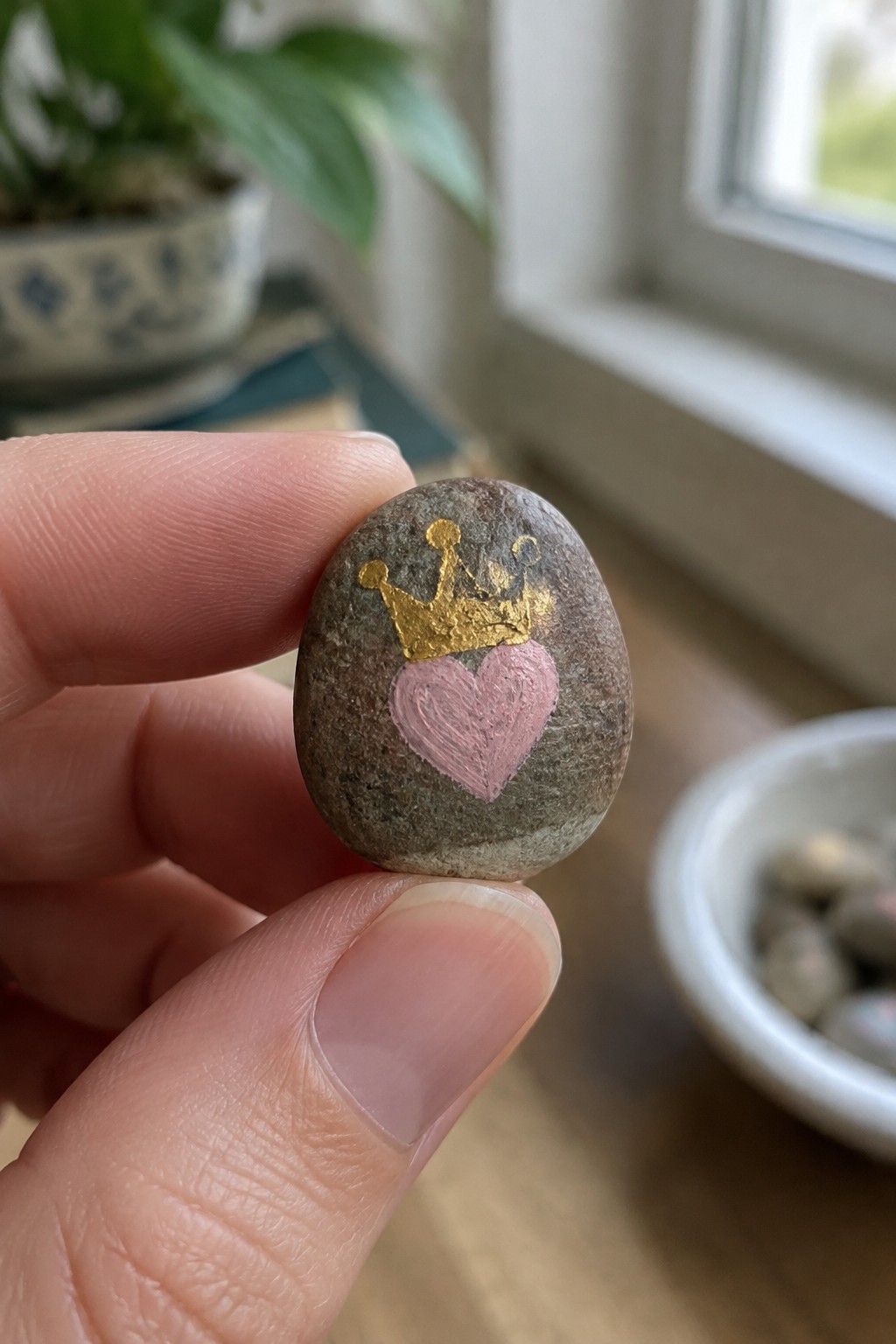 Fingers holding a small stone painted with a pink heart and gold crown