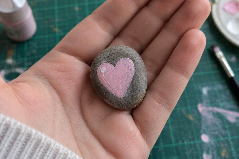 Hand holding gray stone with pink heart amid paint supplies.