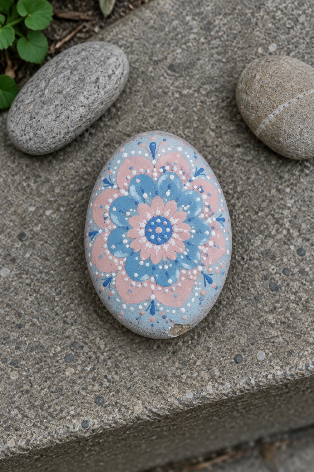 Oval rock with blue and pink floral mandala on gray pavement.