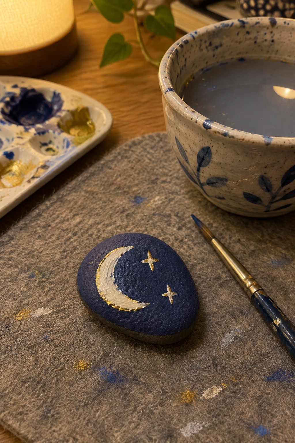 Blue painted stone with gold moon and stars beside paintbrush on felt mat
