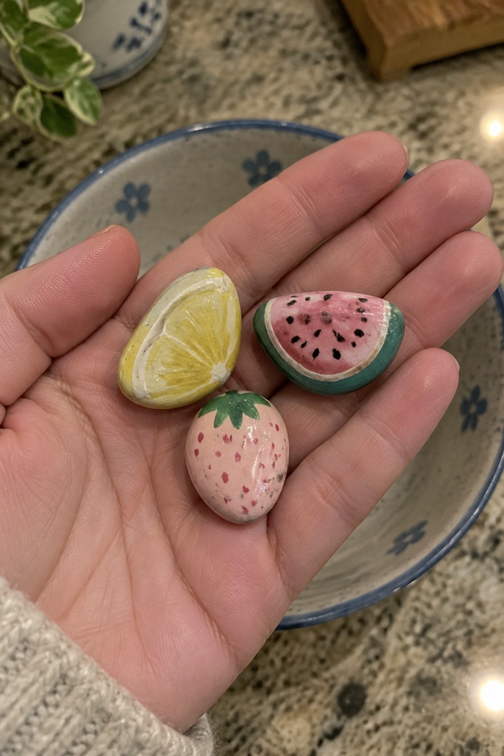 Hand holding three painted rocks resembling lemon, watermelon, and strawberry.