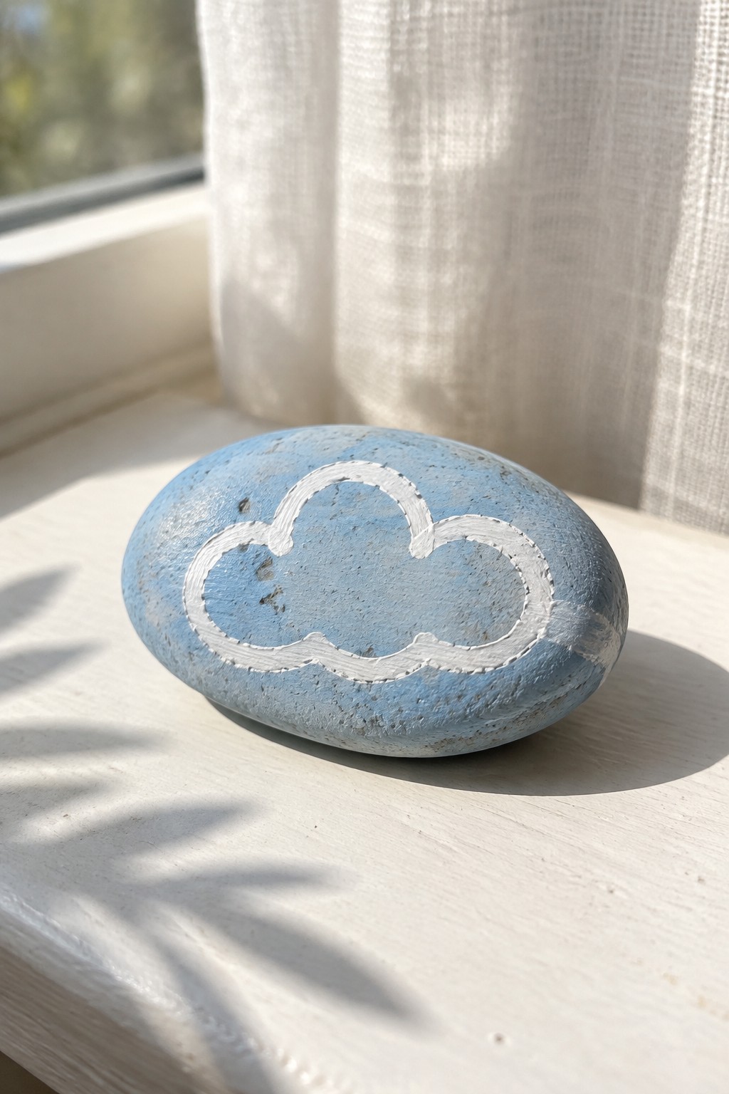 A light blue painted oval rock with a white cloud outline rests on a white windowsill.