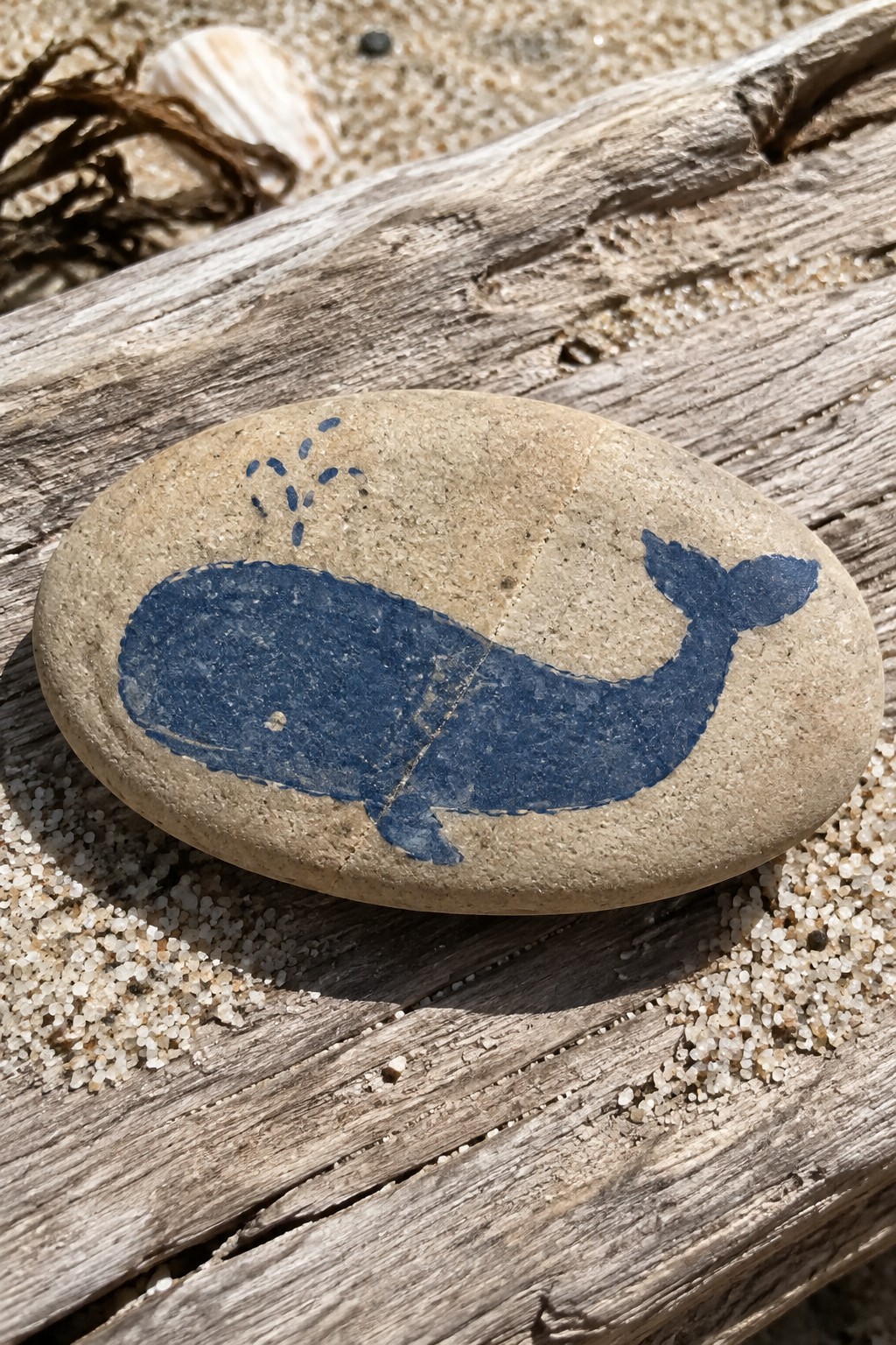 Blue whale painted on smooth oval stone atop weathered driftwood on sand