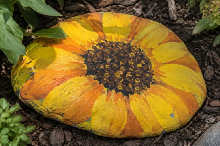 17 Big Rock Painting Ideas With Bold, Oversized Motifs Round painted rock with yellow-orange sunflower petals and dark dotted center on mulch