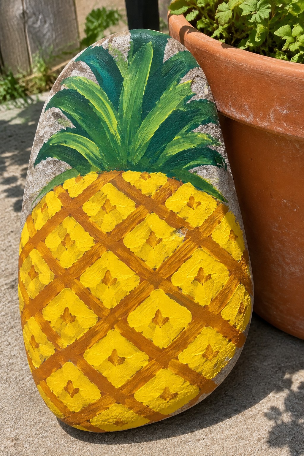 Painted pineapple on oval rock with yellow grid and green leaves beside terracotta pot.
