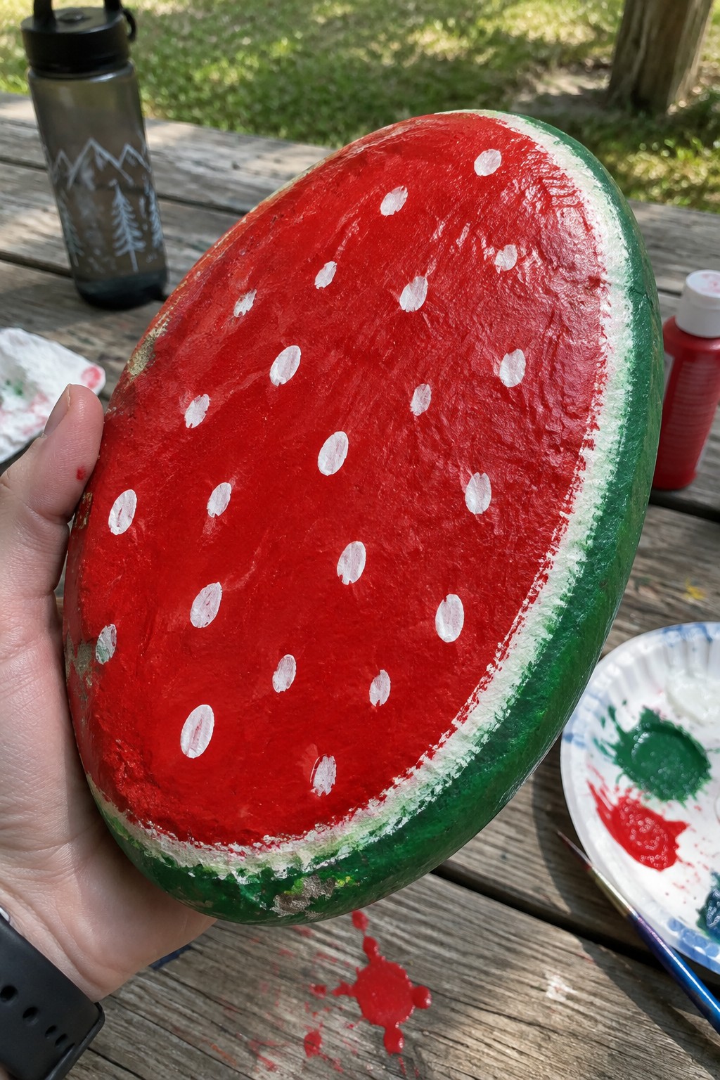 Hand holding red watermelon-painted rock with white seeds on wooden table with supplies.