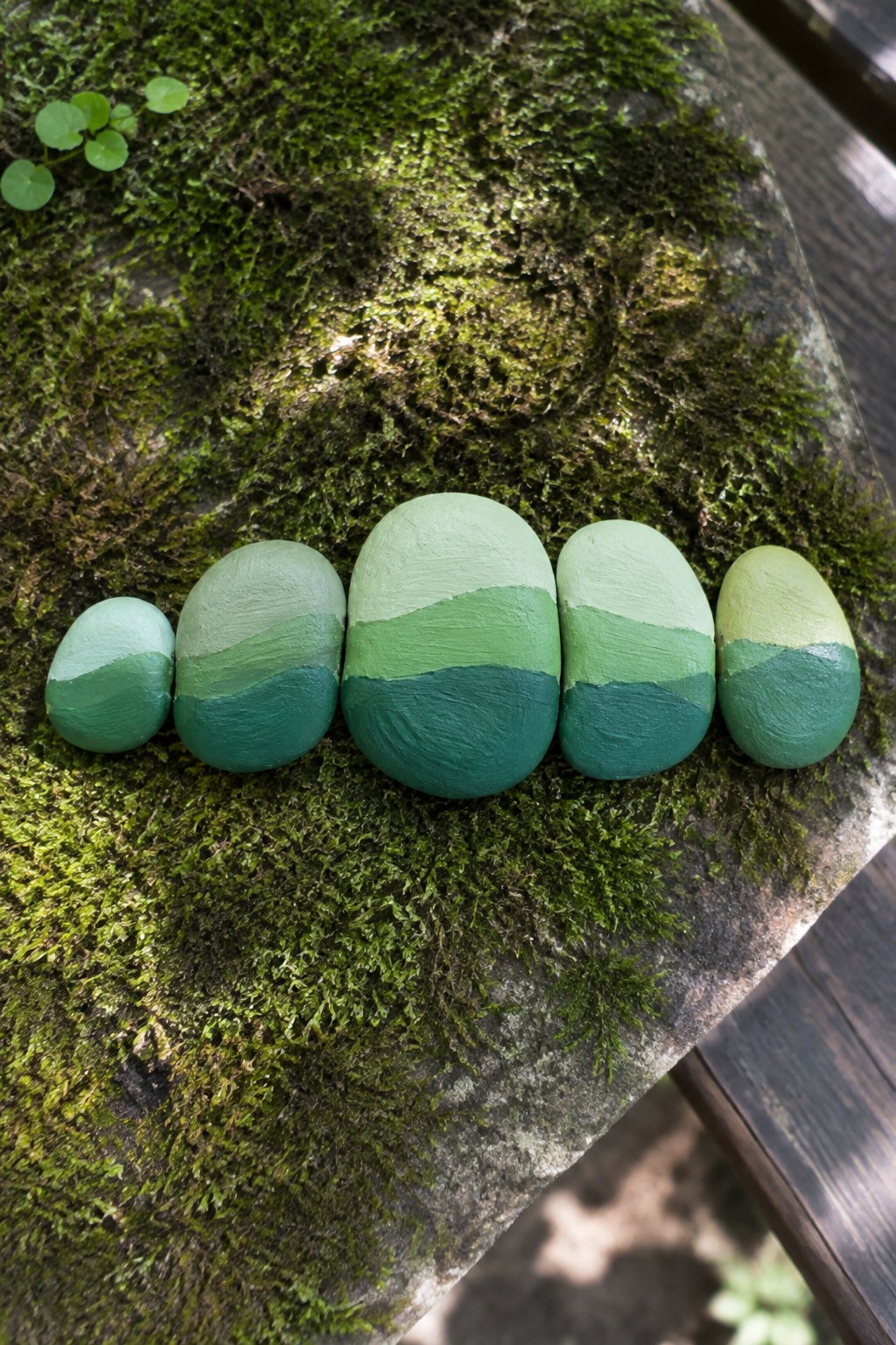 Five green gradient-painted stones lined up on a moss-covered surface