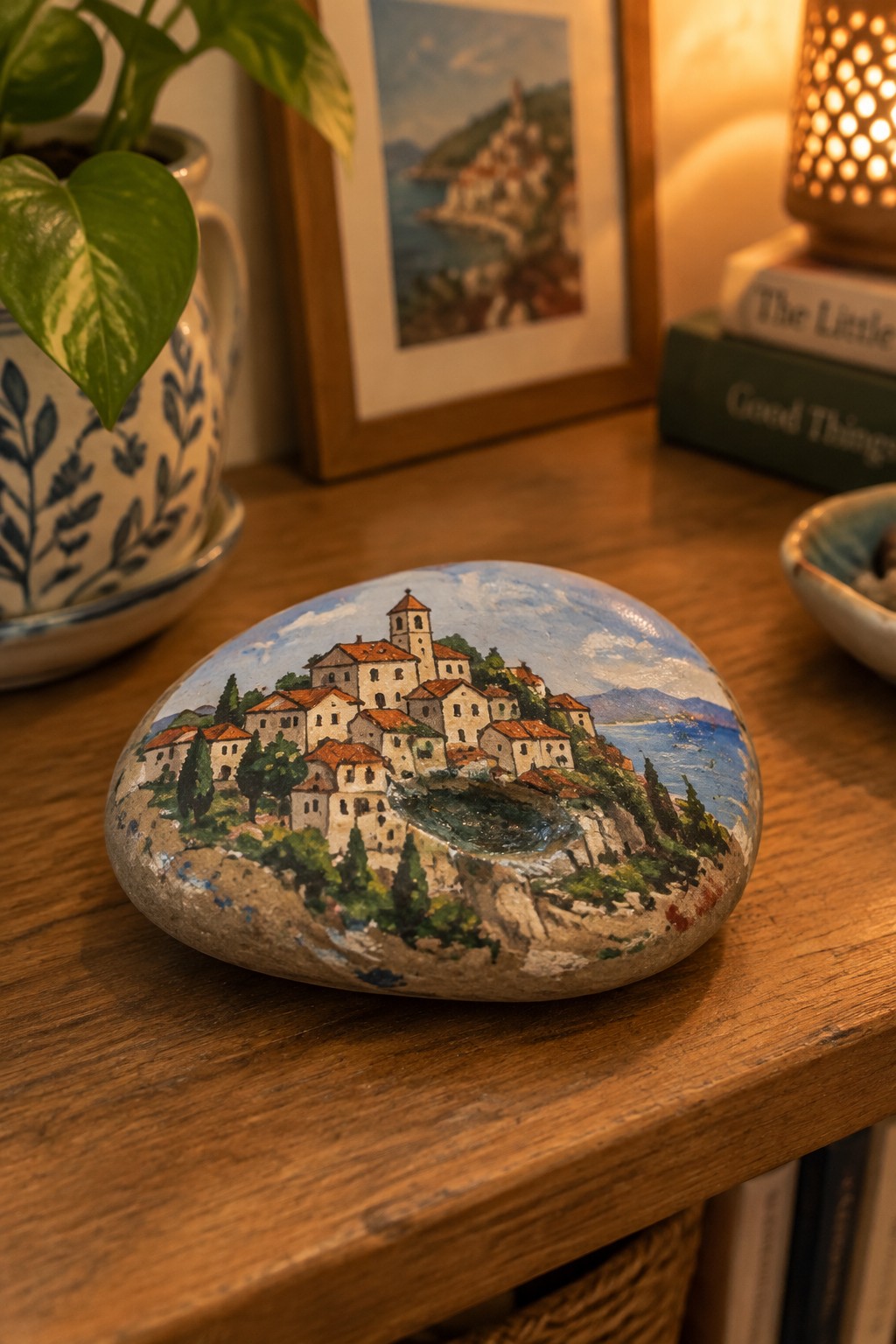 Painted rock showing coastal village with red roofs, church, and sea on wooden table