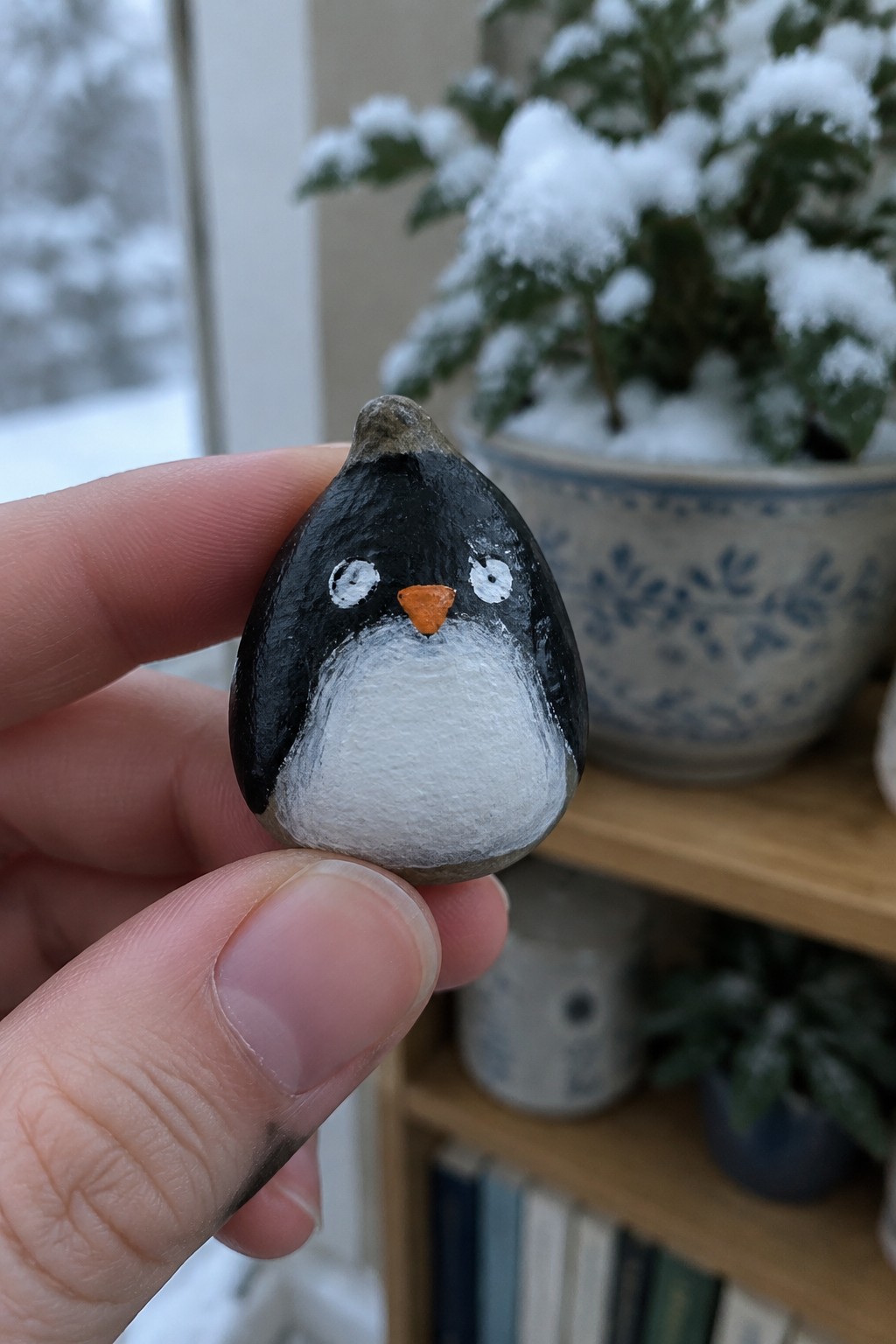 Hand holding small black-and-white painted penguin pebble with orange beak, snowy plant behind
