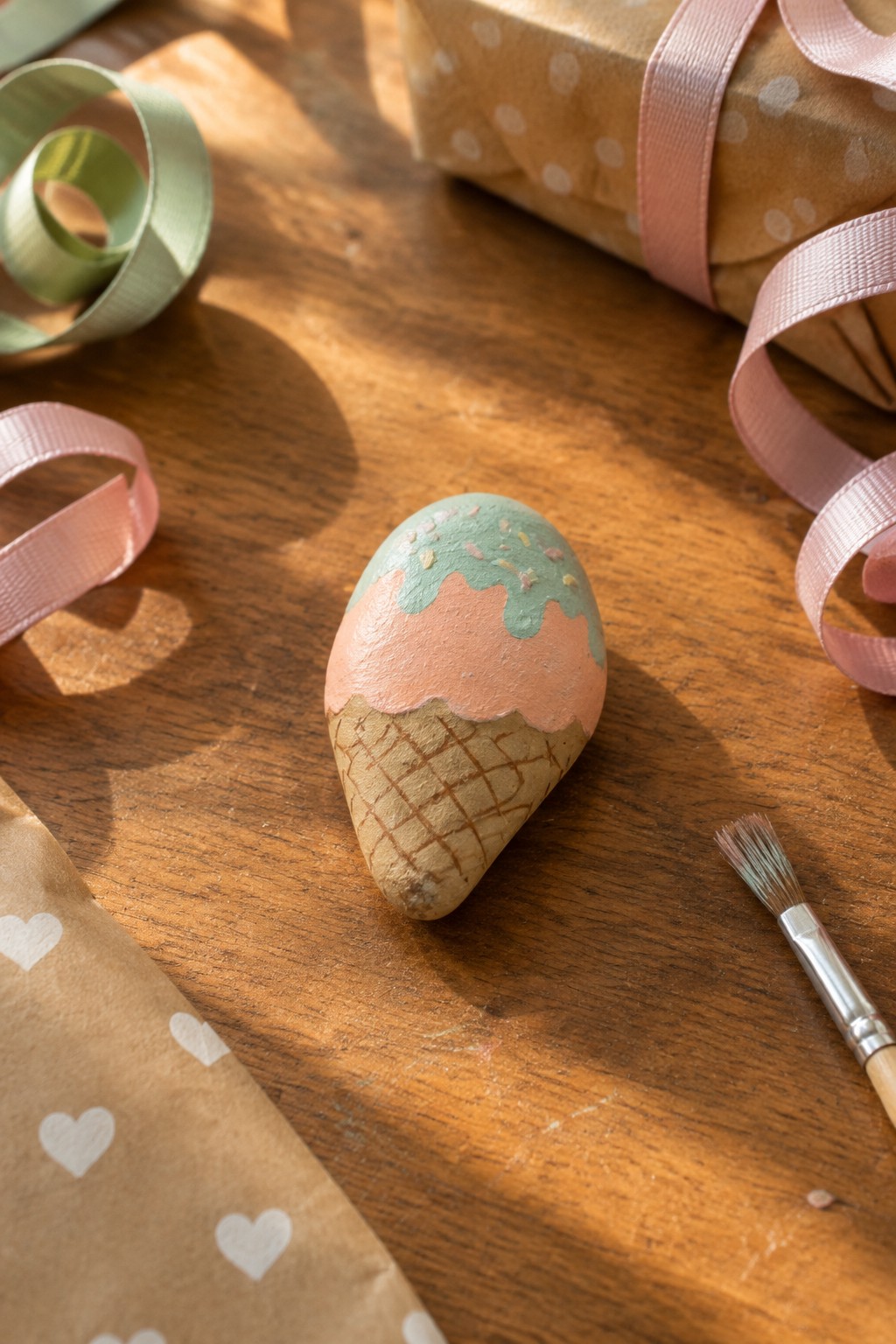 Painted stone ice cream cone on wood with pink ribbons and wrapping paper.