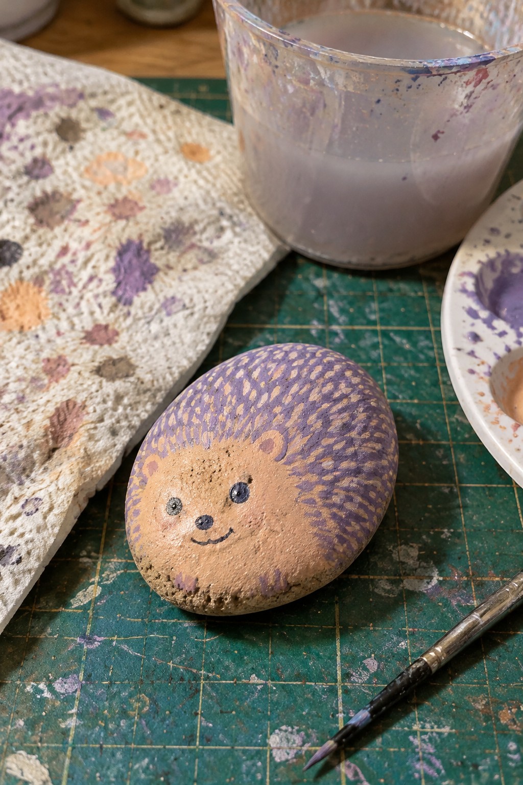 Painted hedgehog rock with purple spines on green mat amid paint supplies