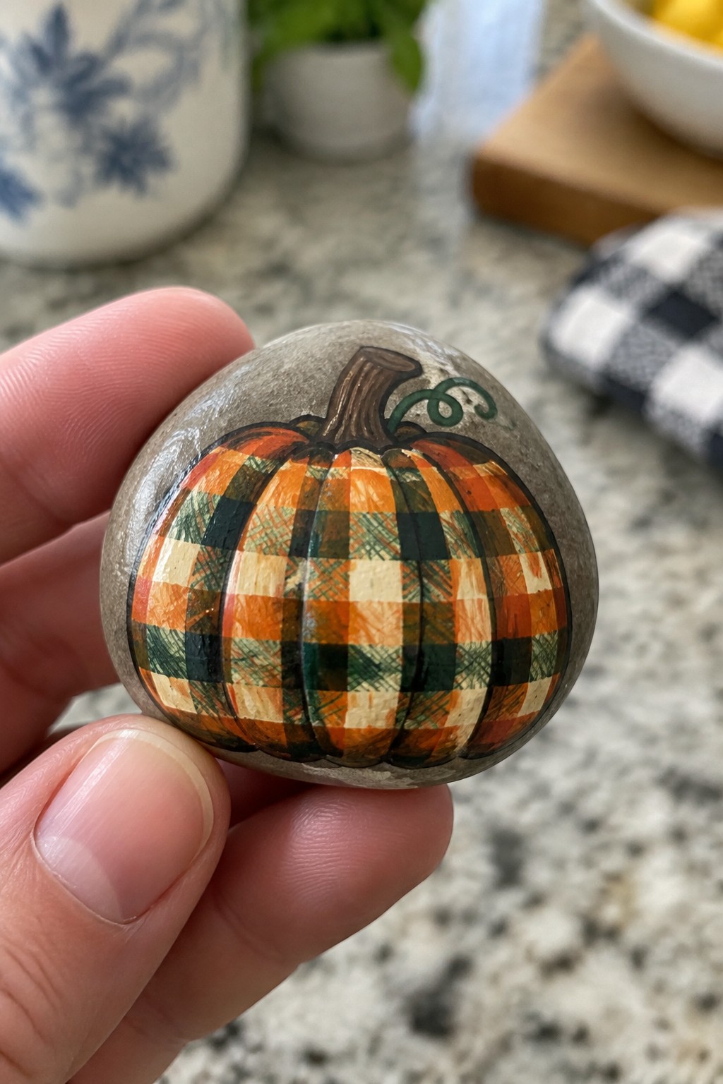 Hand holding plaid pumpkin rock painted orange, green, and beige with brown stem.