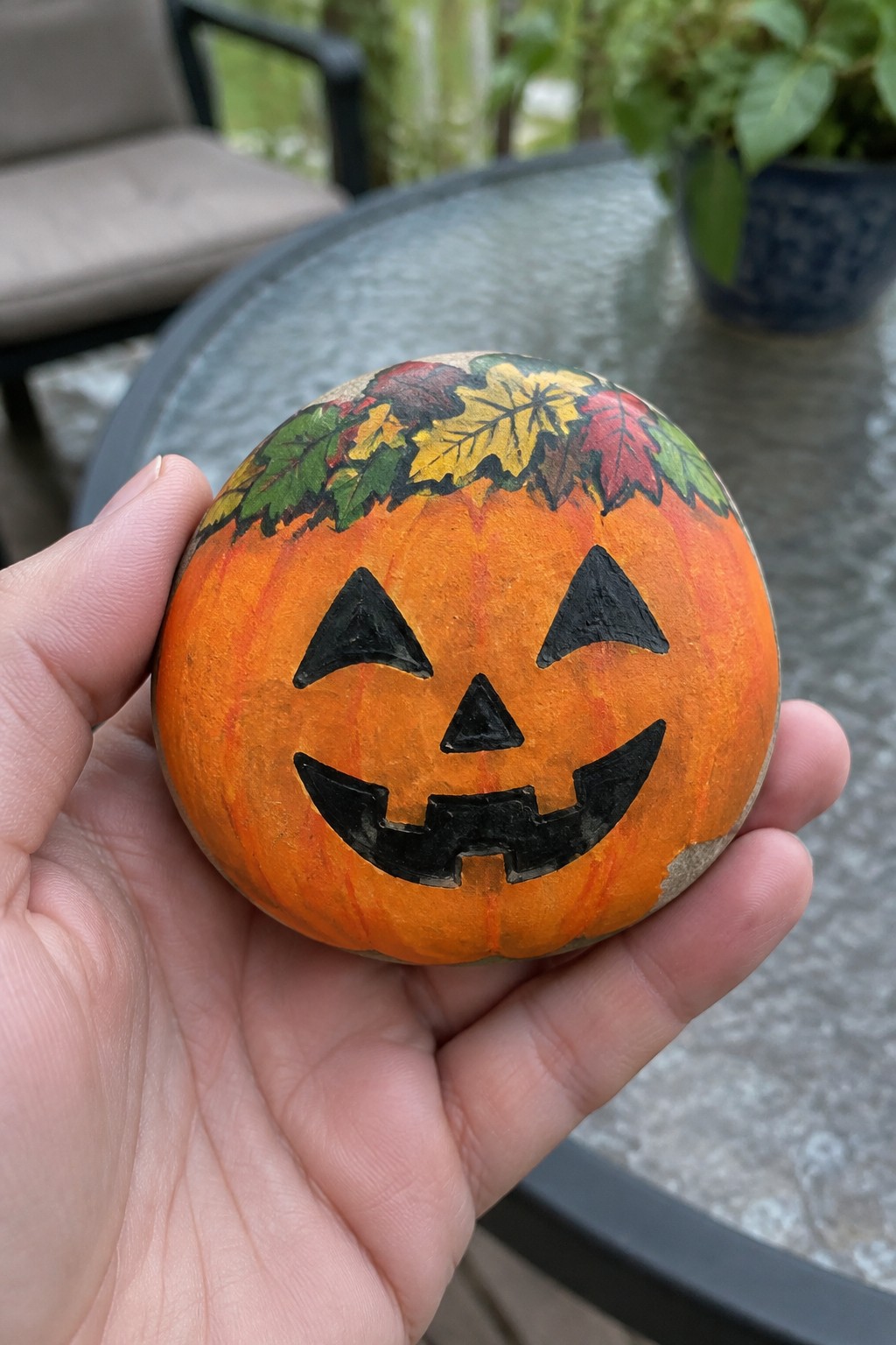 Hand holding painted rock pumpkin with jack-o'-lantern face and autumn leaves