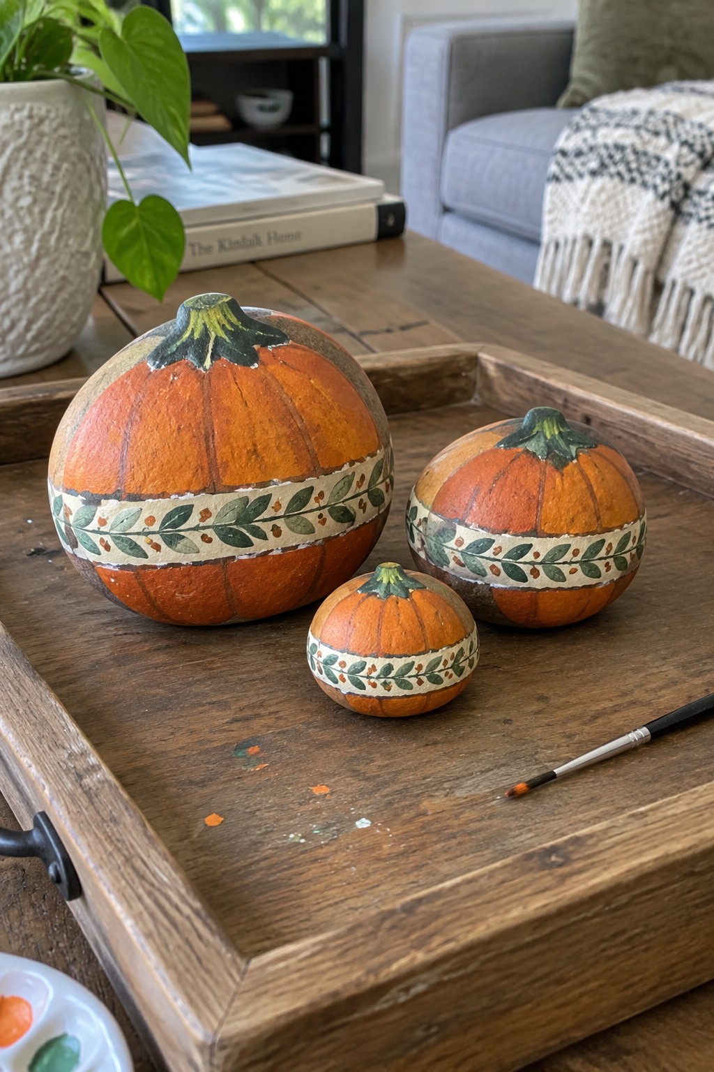 Three painted pumpkins with leaf bands on wooden tray beside paintbrush and palette